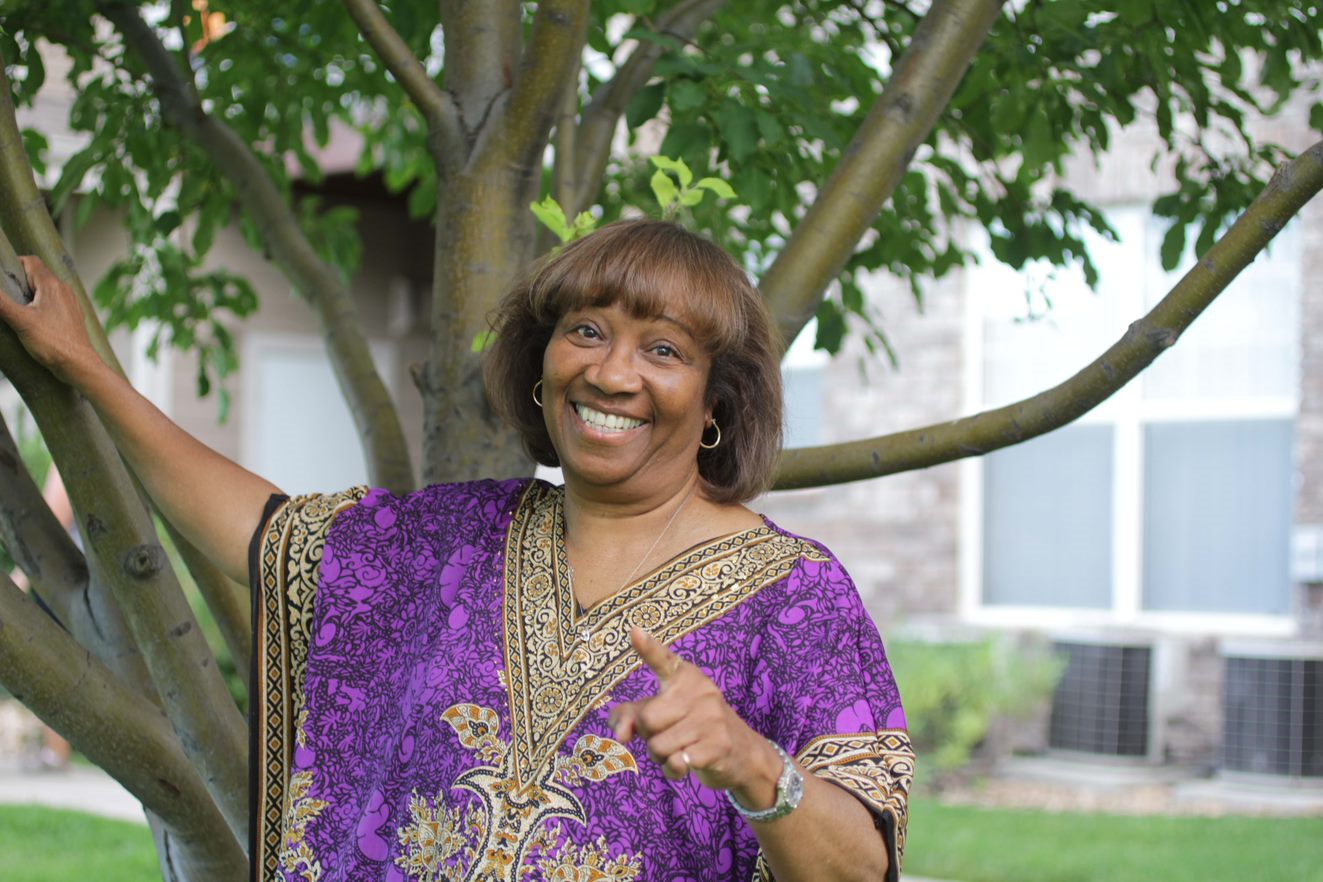 Minister Glenda Robinson pointing to and smiling at the camera in front a tree.