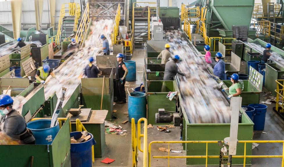 Image of a recycling center, two parallel rows of sorting machines  with workers on eitehr side. 