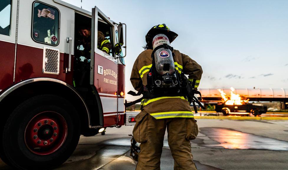 A firefighter in full turnout great stands with their back to the camera. A Boulder Fire-Rescue truck is parked on the left side of the image, and a simulated car fire is on the right side of the image.