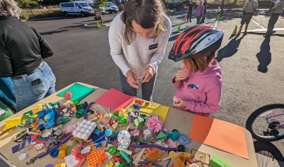 City staff speaks with a child next to a table of miscellaneous toys for a transportation activity.