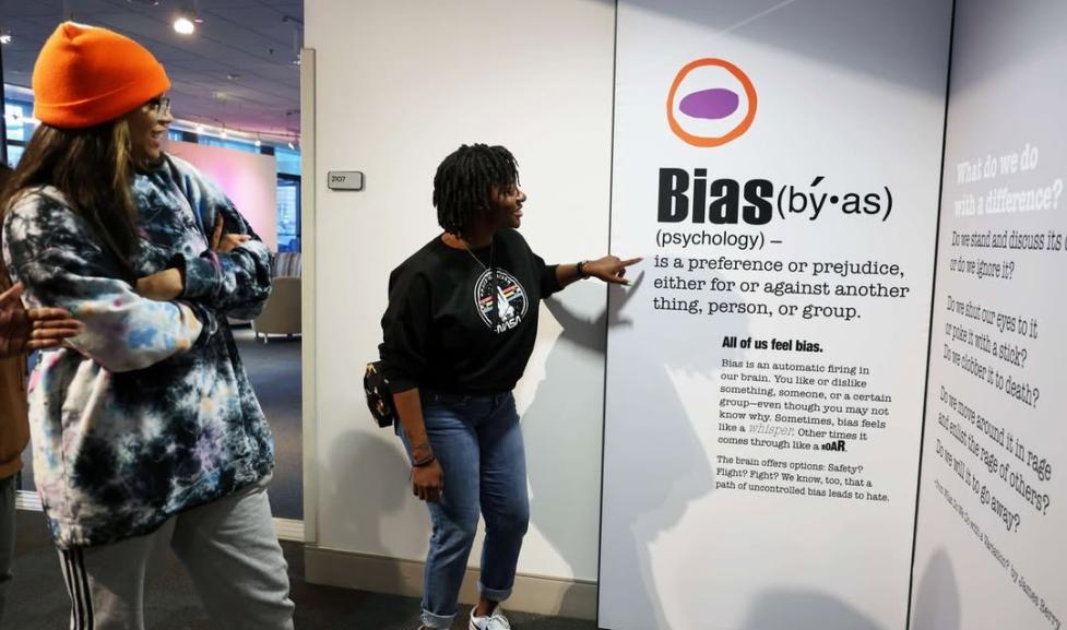 An African American woman wearing jeans and a black t-shirt points to an exhibit wall with 'Bias' and the dictionary definition of bias on it