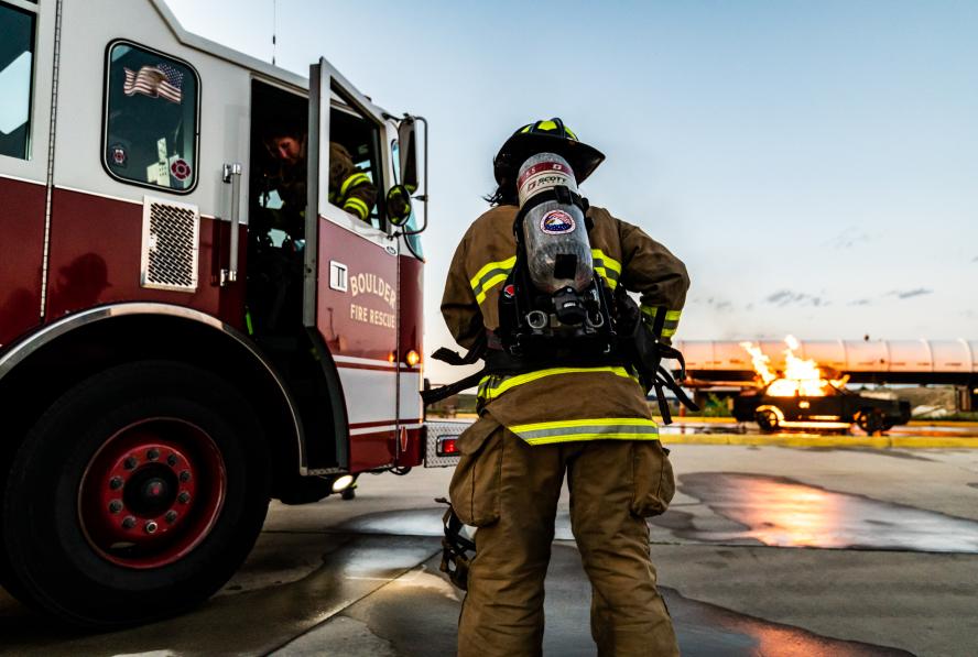 A firefighter in full turnout great stands with their back to the camera. A Boulder Fire-Rescue truck is parked on the left side of the image, and a simulated car fire is on the right side of the image.