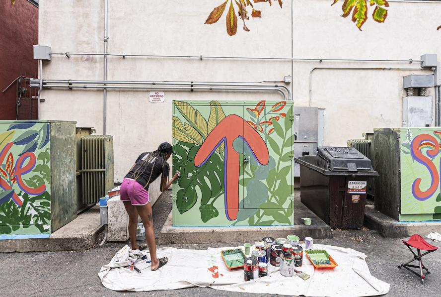 An African American woman with braids tied at the back of her head, wearing a black shirt and pink shorts, bends facing away from the camera to paint a utility box. There are three utility boxes that say 'Take' 'An upwards arrow' and 'Space' or 'take up space'