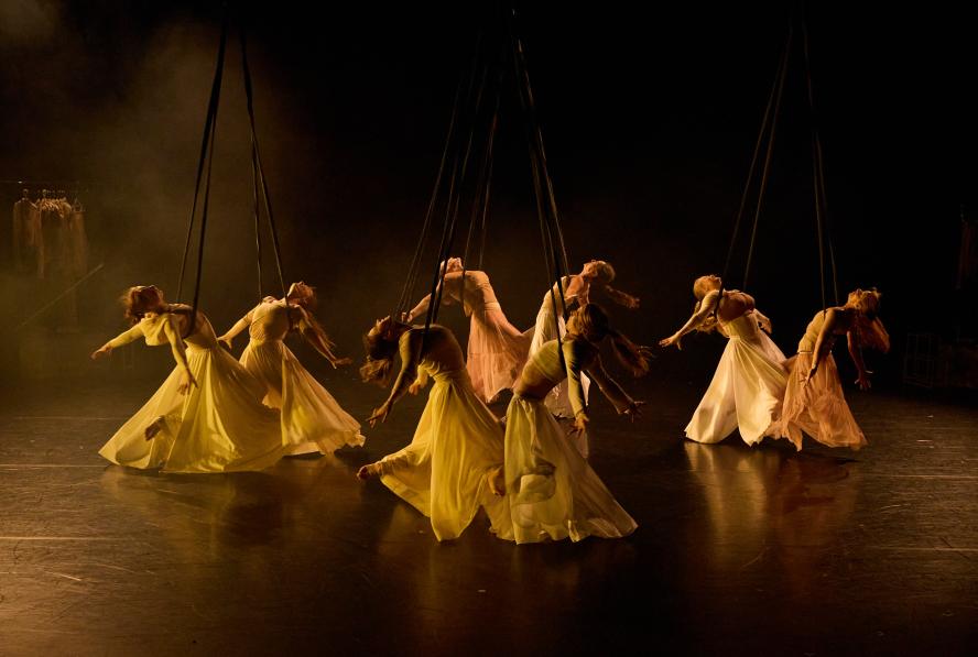 Frequent Flyers dancers perform. A group of seven dancers in white dresses on a dark stage.