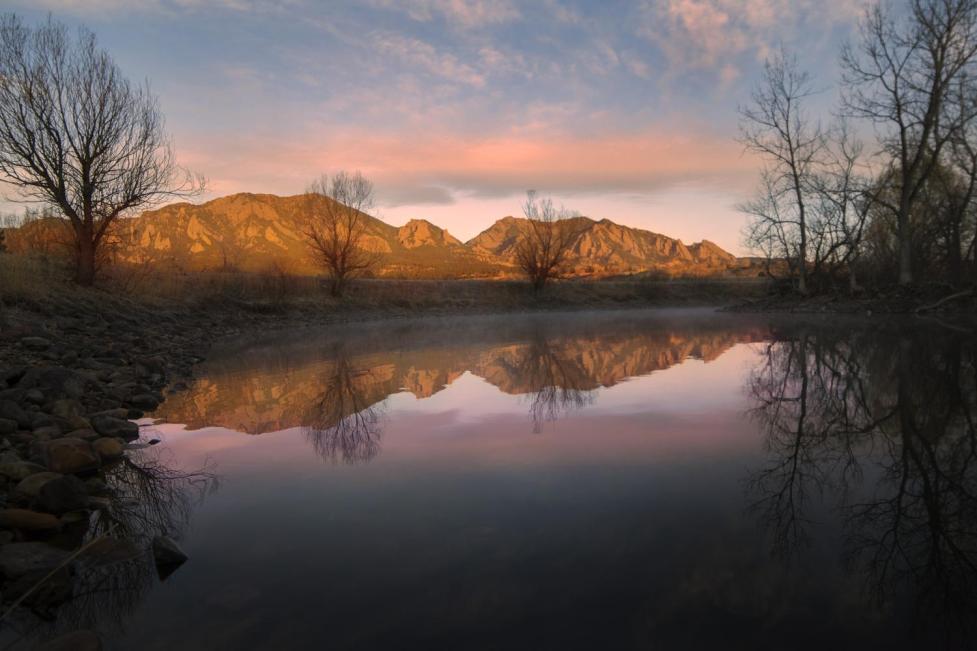 Morning view of the Flatirons lit up by a sunrise with light, pink clouds reflected in still water of the pond at South Boulder Creek trailhead.