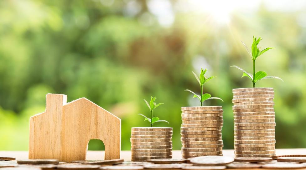 a miniature wooden cutout of a house next to increasingly tall stacks of coins with plant sprouts growing out of the top of the stacks