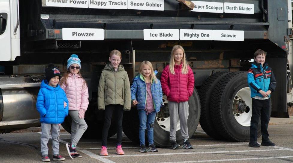 six children standing besides a snowplow with plaques of snowplow names