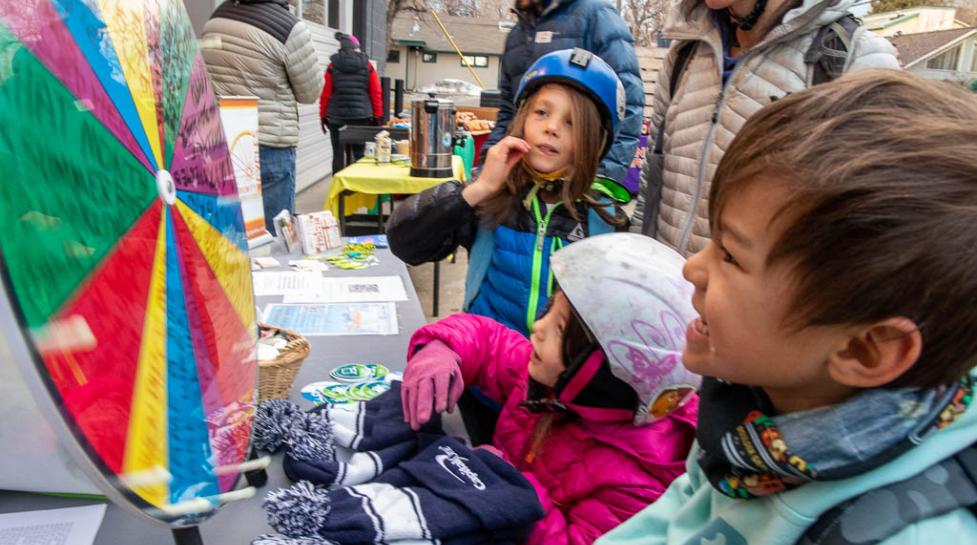 Kids in colorful jackets and helmets outside watch a spinning prize wheel at a table for Winter Bike to Work Day