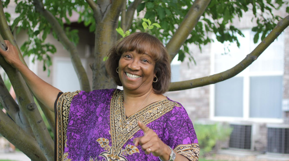 Minister Glenda Robinson pointing to and smiling at the camera in front a tree.