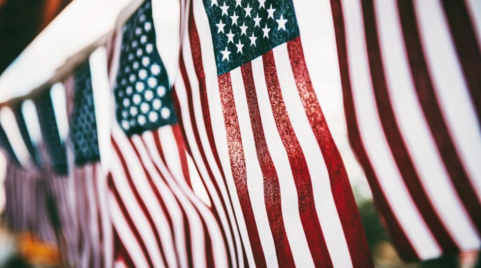 String of American flags hanging vertically with the sun shining through the flags from the background.