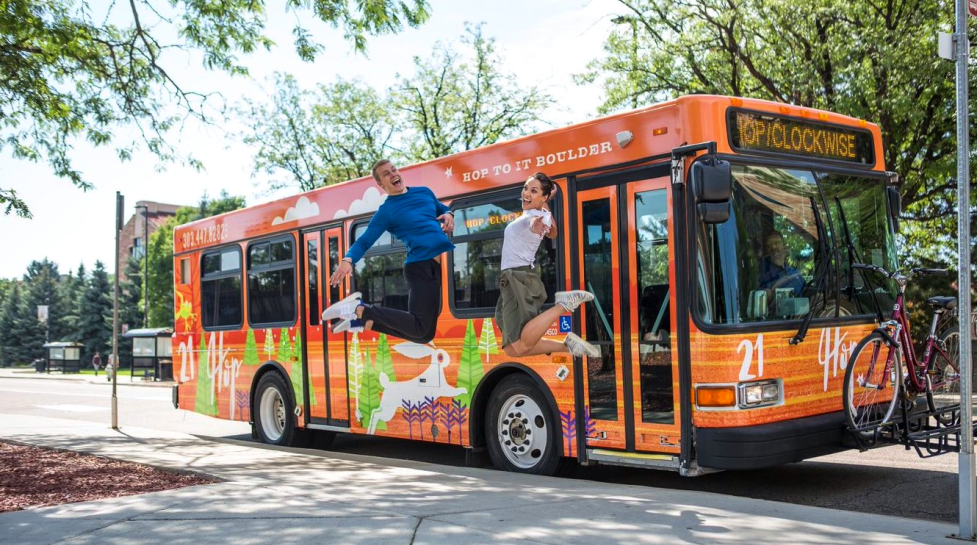 A bright orange HOP bus. Two people are jumping and smiling to the side of the bus. 