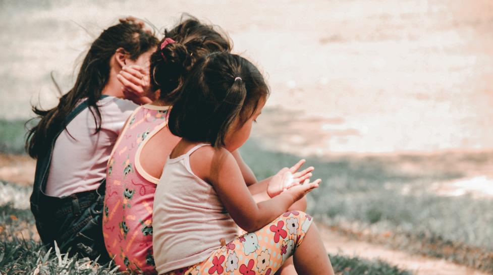 Three children sitting on a grassy hill with their backs to the camera.