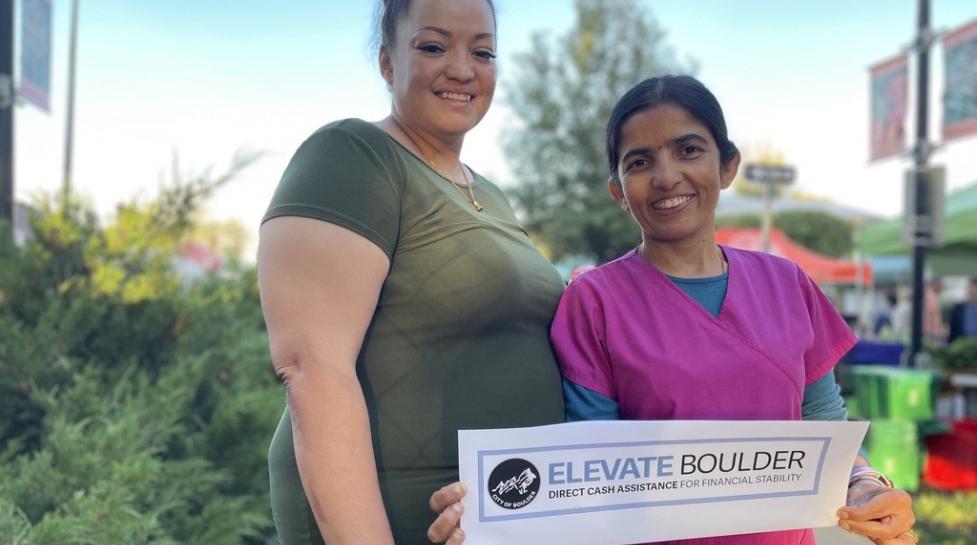 Two people smiling at the camera, the person the right is holding a sign with the Elevate Boulder logo.