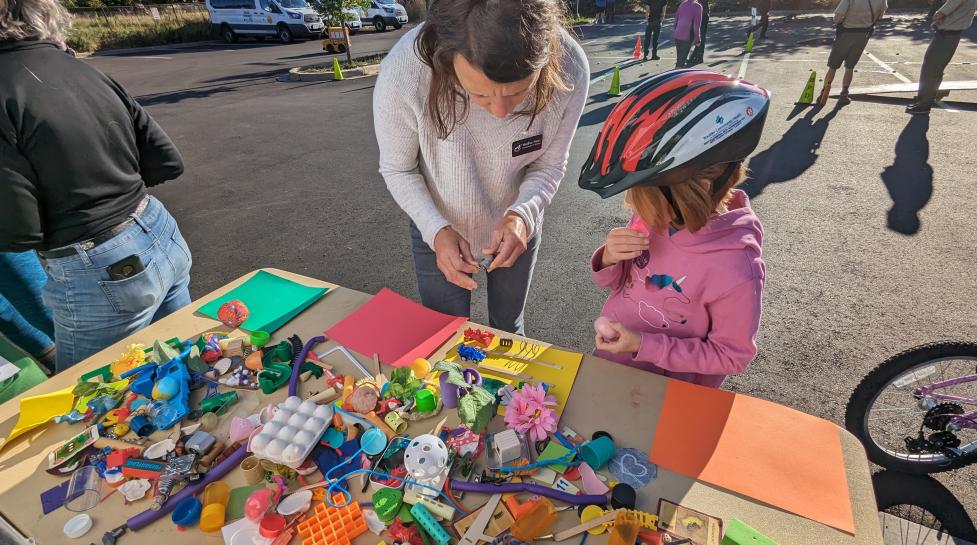 City staff speaks with a child next to a table of miscellaneous toys for a transportation activity.