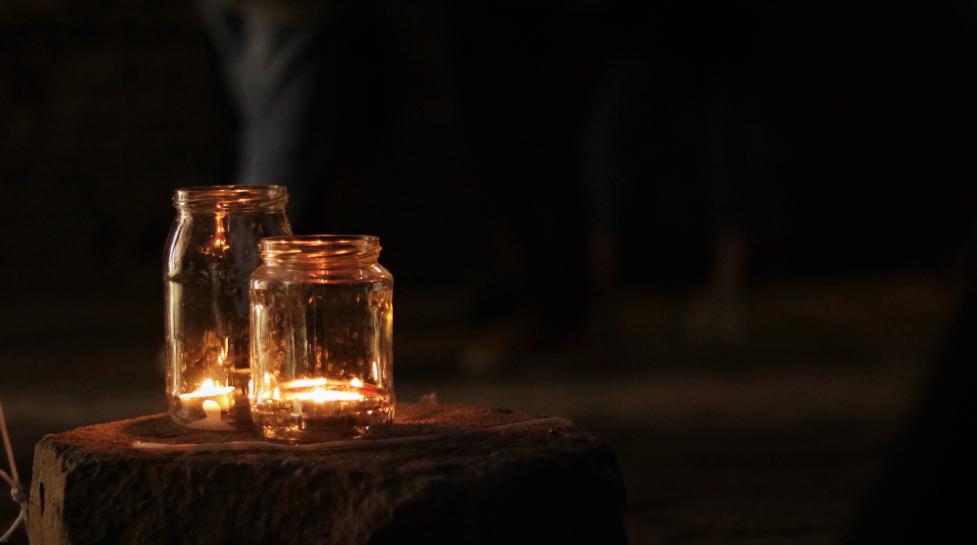 A dark scene with two glass jars lit up with tea light candles sitting on a tree stump 