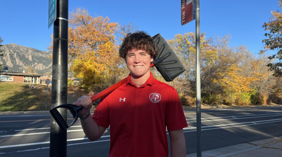 Shovel-a-Stop volunteer Brady Gagliano, a high school student with curly brown hair and a red shirt, smiles while holding a snow shovel in front of a bus stop 