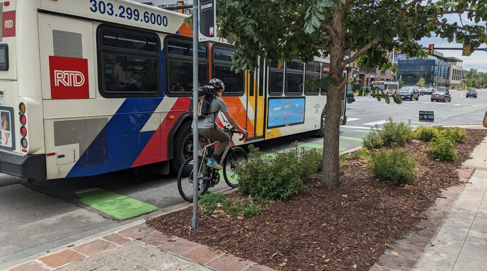 Multimodal travel on north 30th Street, including people biking, driving and taking the bus. A bus is blocking part of the bike lane. 