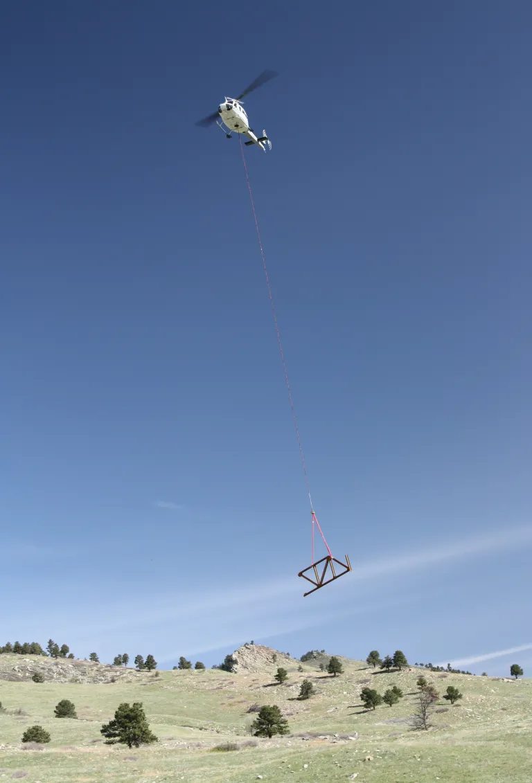 A helicopter flies overhead carrying a piece of metal bridge for North Sky Trail