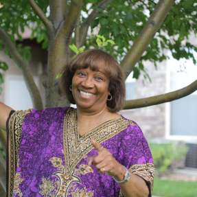 Minister Glenda Robinson pointing to and smiling at the camera in front a tree.