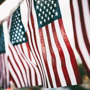 String of American flags hanging vertically with the sun shining through the flags from the background.