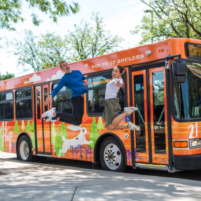 A bright orange HOP bus. Two people are jumping and smiling to the side of the bus. 