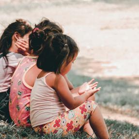 Three children sitting on a grassy hill with their backs to the camera.