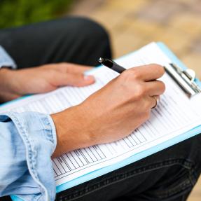 Person sitting down writing on a clipboard that is resting on their knee