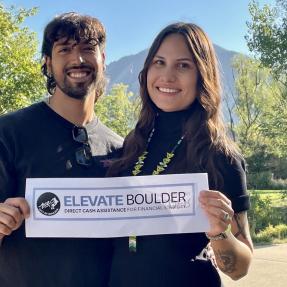 Two people smiling at the camera holding a sign with the Elevate Boulder logo.