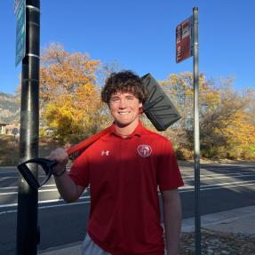 Shovel-a-Stop volunteer Brady Gagliano, a high school student with curly brown hair and a red shirt, smiles while holding a snow shovel in front of a bus stop 