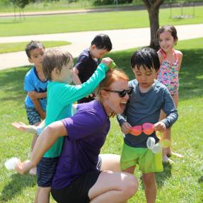 Smiling children pile onto a camp counselor outdoors at one of Boulder’s Kidz Kamp opportunities.