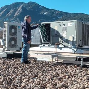 A facilities technician repairing a heating/cooling unit on the rooftop of the Penfield Tate II Municipal Building with the Flatirons in the backdrop.