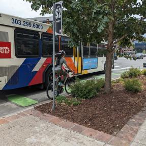 Multimodal travel on north 30th Street, including people biking, driving and taking the bus. A bus is blocking part of the bike lane. 